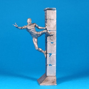 K&ouml;nnte beinhalten: Eine silberne Figur eines Menschen, der eine Wand aus gestapelten Bl&ouml;cken erklimmt. Die Person befindet sich in einer dynamischen Pose mit einem ausgestreckten Bein und einem ausgestreckten Arm.