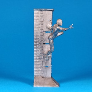 K&ouml;nnte beinhalten: Eine silberne Figur eines Menschen, der eine Ziegelmauer hochklettert. Die Figur ist in einer dynamischen Aktionsstellung mit einem ausgestreckten Arm und dem anderen Arm, der am Ellbogen gebeugt ist, dargestellt. Die Figur steht auf einem kleinen Sockel.