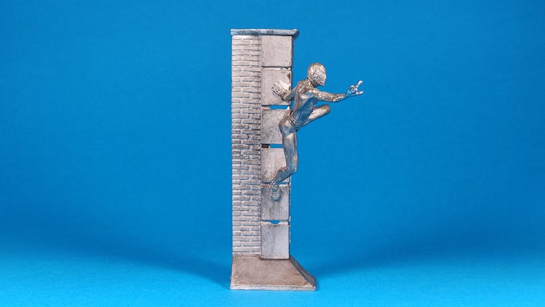 K&ouml;nnte beinhalten: Eine silberne Figur eines Menschen, der eine Ziegelmauer hochklettert. Die Figur ist in einer dynamischen Aktionsstellung mit einem ausgestreckten Arm und dem anderen Arm, der am Ellbogen gebeugt ist, dargestellt. Die Figur steht auf einem kleinen Sockel.