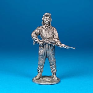 May include: A silver-colored metal figurine of a soldier wearing a hat and holding a rifle. The figurine is standing on a circular base.