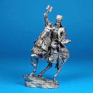 Puede incluir: Figurilla detallada de color plateado de un guerrero montado, sosteniendo un arma en alto. La figura está a caballo, con armadura intrincada y un sombrero alto. La base es una formación rocosa sobre un fondo azul.