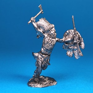 May include: A silver-colored figurine of a Native American warrior in a dynamic pose. The figure is holding a tomahawk overhead and a rifle and shield in the other hand. The warrior is wearing a headdress and traditional clothing.