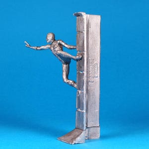 K&ouml;nnte beinhalten: Eine silberne Figur eines Menschen, der eine Wand hochklettert. Die Figur ist in einer dynamischen Aktionsstellung mit einem ausgestreckten Bein und dem anderen Bein, das im Knie gebeugt ist, dargestellt. Die Wand ist strukturiert und hat eine erhabene Inschrift mit der Aufschrift "ANDERSEN GARAGE".