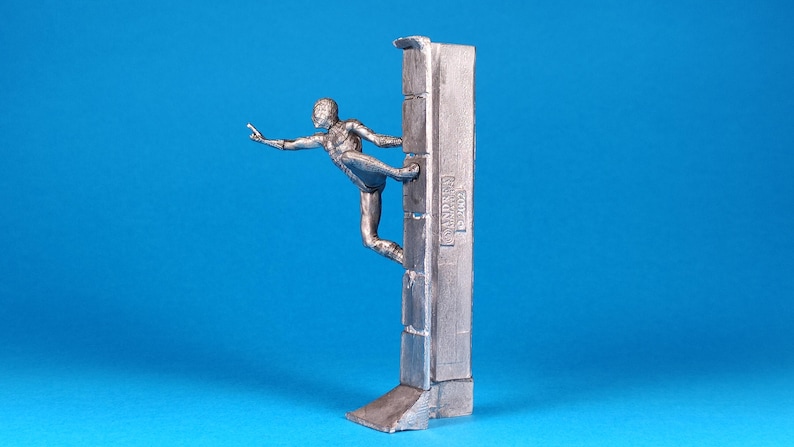 K&ouml;nnte beinhalten: Eine silberne Figur eines Menschen, der eine Wand hochklettert. Die Figur ist in einer dynamischen Aktionsstellung mit einem ausgestreckten Bein und dem anderen Bein, das im Knie gebeugt ist, dargestellt. Die Wand ist strukturiert und hat eine erhabene Inschrift mit der Aufschrift "ANDERSEN GARAGE".