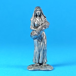 May include: A silver-colored figurine of a woman in a flowing gown, holding a basket and wheat stalks. The figure stands on a textured base against a solid blue background. The statue has long hair and is detailed.