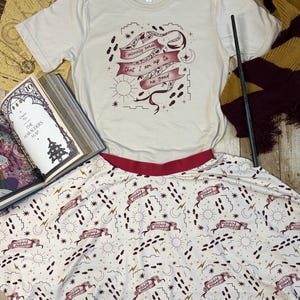 May include: A cream-colored t-shirt with the text "I solemnly swear that I am up to no good" and a matching skirt. The skirt has a white background with a repeating pattern of footprints, stars, and banners that say "Mischief Managed". A wand and a book are also in the image.