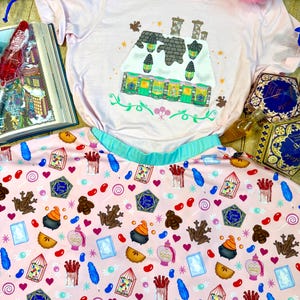 May include: A light pink t-shirt and skirt set. The t-shirt features a colorful illustration of a building with green and yellow accents. The skirt has a light pink background with various candy and magical-themed graphics, including chocolate frogs and jelly beans.
