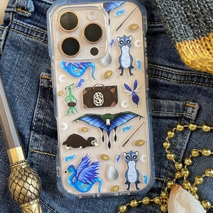Puede incluir: Una funda transparente para teléfono con un diseño colorido que presenta criaturas mágicas, incluyendo un fénix azul y morado, un Niffler marrón y blanco, un  Swooping Evil azul y verde, y un  Thunderbird azul y blanco. La funda también presenta una maleta con un símbolo S dorado, una varita mágica y una moneda de oro.