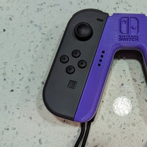 Peut inclure: Une manette Nintendo Switch Joy-Con de couleur violette et noire avec le logo Nintendo Switch sur le devant.