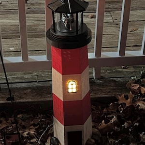 May include: A solar-powered lighthouse with a red and white brick design. The lighthouse has a black top and a small window that emits a warm glow. It is surrounded by leaves and small rocks.