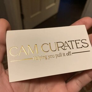May include: A white business card with gold lettering that reads "CAM CURATES" and "Helping you pull it off."
