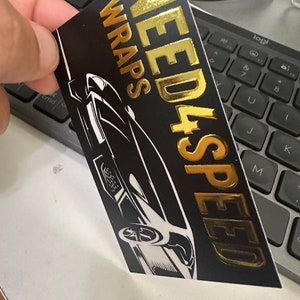 May include: Black and gold sticker with the text "NEED 4 SPEED WRAPS" in gold lettering. A black and white silhouette of a sports car is in the background.