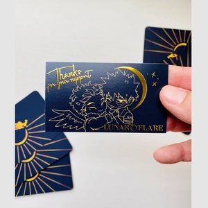 May include: A gold foil thank you card with a black background. The card features a cartoon illustration of two characters with a crescent moon and stars. The text on the card reads "Thanks for your support" and "LUNAROFLARE".