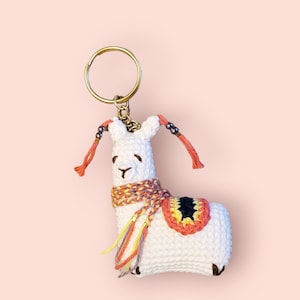 Peut inclure: Porte-clés en forme de lama en crochet blanc avec une écharpe rose, jaune et violette. Le lama a des yeux noirs et un nez noir. Le porte-clés a un anneau en métal doré.