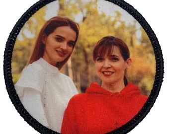 May include: Two women are standing in a forest. The woman on the left is wearing a white shirt and the woman on the right is wearing a red hoodie. They are both smiling.