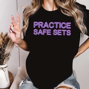 May include: A black t-shirt with the text "PRACTICE SAFE SETS" in purple neon lettering.