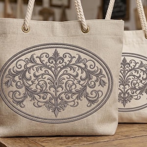 Puede incluir: Dos bolsas tote beige con asas de cuerda. Cada bolsa presenta un diseño ovalado gris con un patrón floral y de volutas detallado. Las bolsas están hechas de una tela texturizada y se exhiben sobre una superficie de madera.