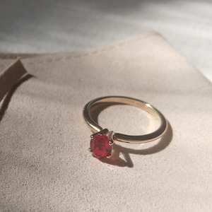 Puede incluir: Un anillo de oro con una gema roja y redonda. El anillo está sobre una superficie clara, con una sombra proyectada por la luz solar. La banda del anillo es delgada y la gema es el foco.