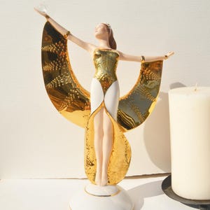 Puede incluir: Una figura de porcelana con los brazos extendidos, vestida con un traje dorado y blanco con grandes paneles dorados reflectantes en forma de alas. La figura sostiene una pequeña esfera transparente y se apoya sobre una base circular.