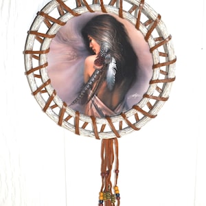 May include: Round dreamcatcher featuring a portrait of a woman with long dark hair and feathers. The frame is white with brown leather accents. Brown leather strands with beads hang from the bottom.