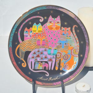 May include: A black ceramic plate with a gold rim features a colourful illustration of a group of cats. The cats are painted in various shades of pink, orange, blue, green, and yellow. The plate is signed "Anne Burch" in the bottom right corner.
