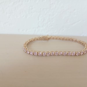 Könnte beinhalten: Ein goldfarbenes Tennisarmband mit rosa Edelsteinen. Das Armband liegt auf einer hellen Oberfläche. Die Edelsteine sind quadratisch geschliffen und eng aneinander gesetzt, wodurch eine durchgehende Linie entsteht.