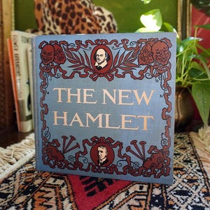 May include: A hardcover book titled "The New Hamlet" with a light blue cover. The cover features decorative red and brown accents, including portraits and theatrical masks. The book is displayed on a patterned rug.