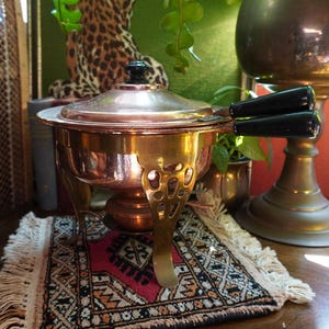 Peut inclure: Un chafing dish cuivré avec une poignée et un bouton de couvercle noirs, posé sur un support décoratif. Le plat repose sur un petit tapis avec un motif rouge et noir. L'arrière-plan comprend un imprimé léopard et de la verdure.