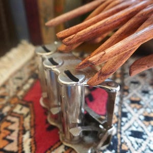 May include: A silver-toned metal coin dispenser featuring three vertical cylindrical tubes with individual slots at the top for inserting coins, accompanied by a wooden hand-shaped sculpture with articulated fingers resting above the device.