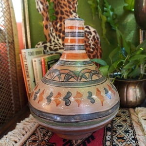 May include: A hand-painted ceramic vase with a bulbous body and a narrow neck. The vase features a tan base with orange, black, and blue decorative patterns. The neck has horizontal stripes in orange, black, and tan. The vase is a decorative home accent.
