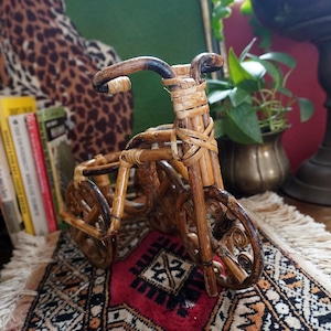 May include: A miniature decorative bicycle made of woven bamboo. The bicycle has a curved handlebar and spoked wheels. It sits on a small rug with a red and black geometric pattern. The background includes books and a green wall.