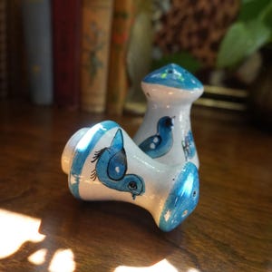 Puede incluir: Dos saleros y pimenteros de cerámica con base blanca y motivos de pájaros pintados a mano en azul. Uno está de pie y el otro tumbado. Ambos tienen forma de reloj de arena con bordes pintados en azul y pequeños orificios en la parte superior.