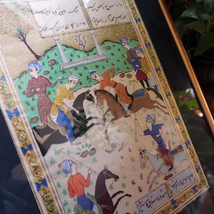 May include: A framed miniature painting depicting a polo match with multiple figures on horseback, wearing colorful tunics and turbans. The scene includes green landscape elements, architectural pillars, and Persian script in the top and bottom borders.