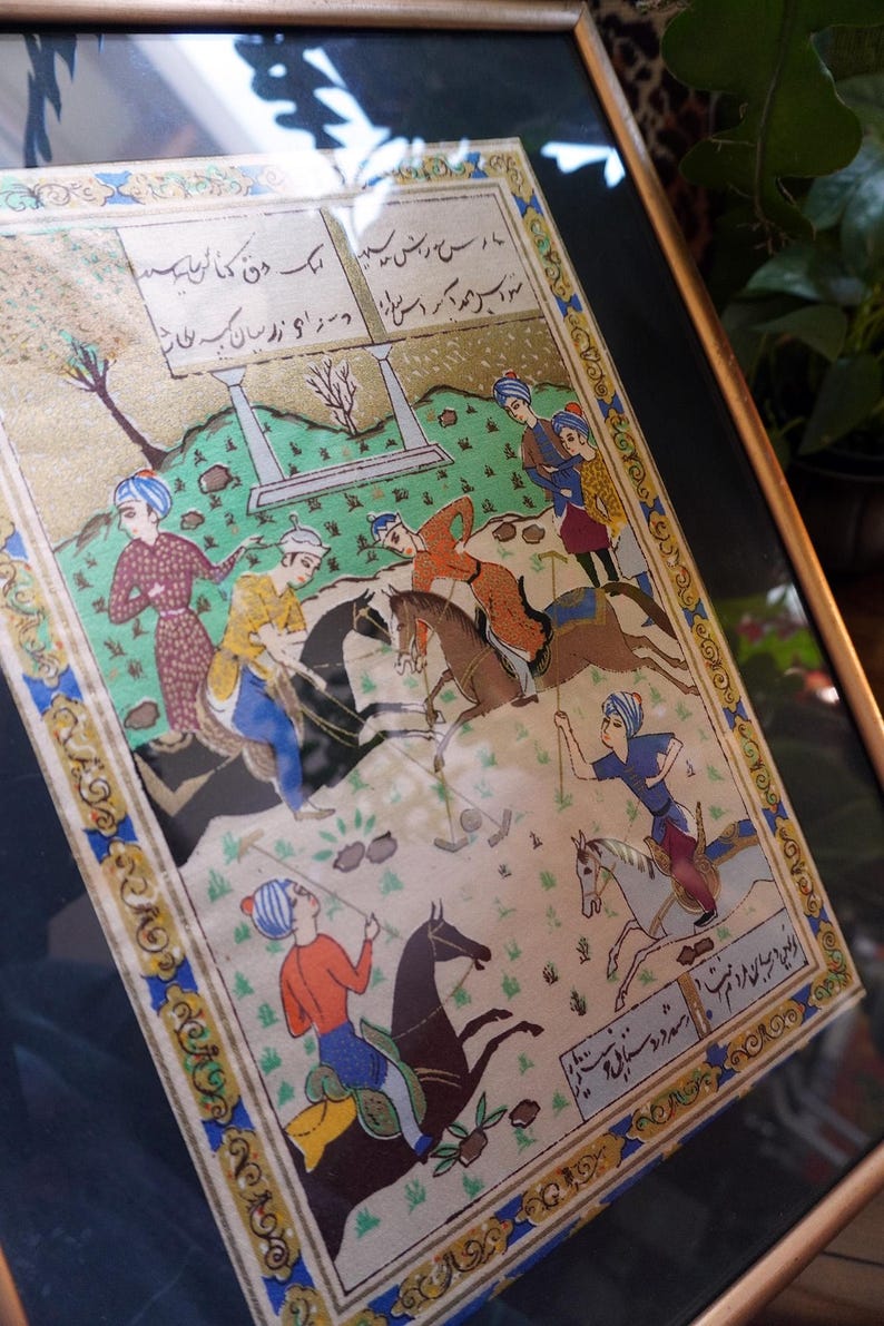 May include: A framed miniature painting depicting a polo match with multiple figures on horseback, wearing colorful tunics and turbans. The scene includes green landscape elements, architectural pillars, and Persian script in the top and bottom borders.