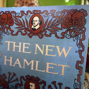 May include: A vintage book titled "The New Hamlet" with a light blue cover. The cover features decorative brown accents, including portraits and theatrical masks. The title is in gold lettering.