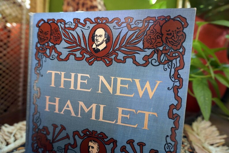May include: A vintage book titled "The New Hamlet" with a light blue cover. The cover features decorative brown accents, including portraits and theatrical masks. The title is in gold lettering.