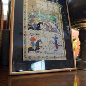 May include: A framed miniature painting depicting a polo match with four riders on horseback, wearing colorful tunics and turbans. The scene includes Persian script at the top and bottom, set against a green landscape with a decorative blue and gold border.