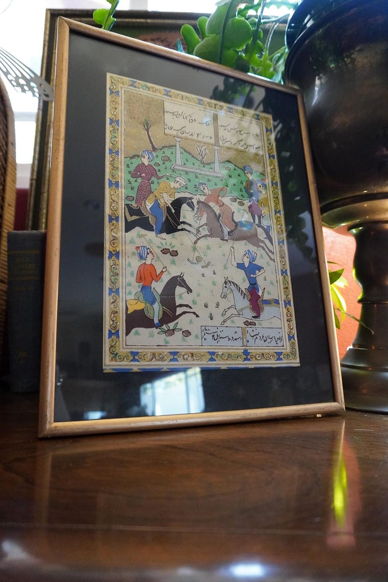 May include: A framed miniature painting depicting a polo match with four riders on horseback, wearing colorful tunics and turbans. The scene includes Persian script at the top and bottom, set against a green landscape with a decorative blue and gold border.