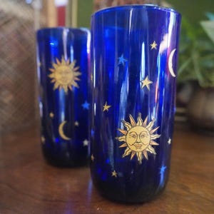 May include: Two cylindrical drinking glasses made of deep cobalt blue glass. Each glass features a printed design of a golden sun with a human face, surrounded by various small yellow and blue stars and a crescent moon scattered across the surface.