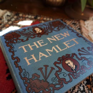 May include: A hardcover book titled "THE NEW HAMLET" with a light blue cover. The cover features ornate brown illustrations and portraits, with the title in large, light-colored letters. The book rests on a patterned rug.