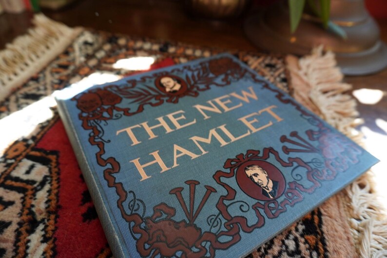 May include: A hardcover book titled "THE NEW HAMLET" with a light blue cover. The cover features ornate brown illustrations and portraits, with the title in large, light-colored letters. The book rests on a patterned rug.
