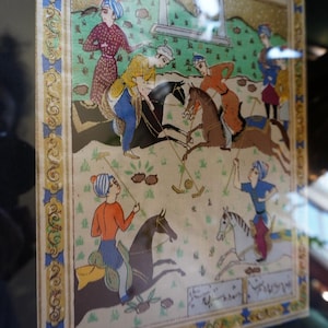 May include: A rectangular painting depicting a polo match with four riders on horseback, each wearing colorful traditional attire and turbans. The scene includes green grass, brown horses, and decorative borders with Persian script at the top and bottom.