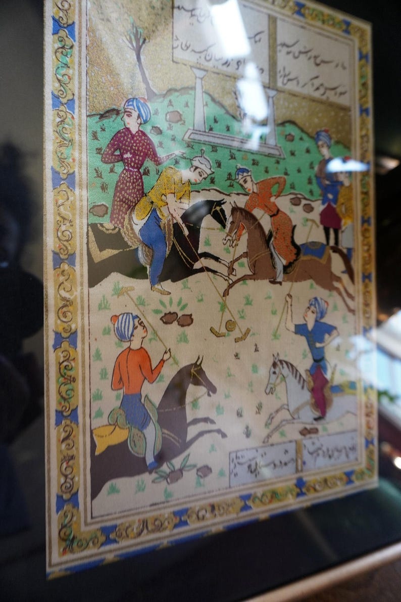 May include: A rectangular painting depicting a polo match with four riders on horseback, each wearing colorful traditional attire and turbans. The scene includes green grass, brown horses, and decorative borders with Persian script at the top and bottom.