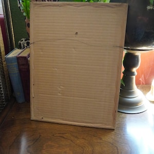 May include: A rectangular picture frame viewed from the back, featuring a light brown corrugated cardboard backing. A thin, dark metal hanging wire is attached to the top section of the frame, spanning horizontally across the center of the cardboard.