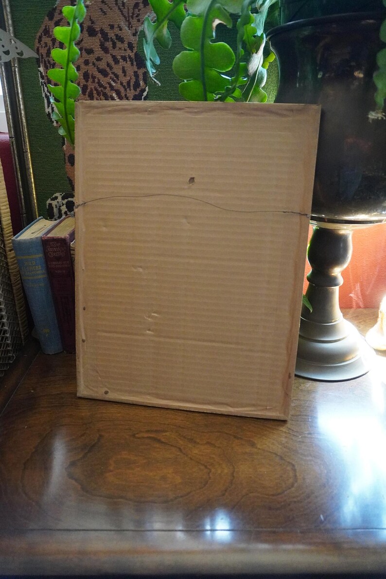 May include: A rectangular picture frame viewed from the back, featuring a light brown corrugated cardboard backing. A thin, dark metal hanging wire is attached to the top section of the frame, spanning horizontally across the center of the cardboard.