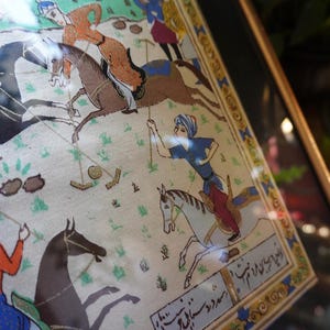 May include: A framed miniature painting on fabric depicting a polo match with riders on horseback. The scene features figures in colorful traditional attire, horses in motion, and a polo ball on the ground. The artwork includes decorative borders and script.