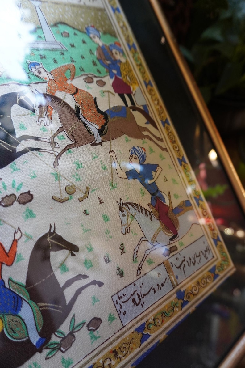 May include: A framed miniature painting on fabric depicting a polo match with riders on horseback. The scene features figures in colorful traditional attire, horses in motion, and a polo ball on the ground. The artwork includes decorative borders and script.