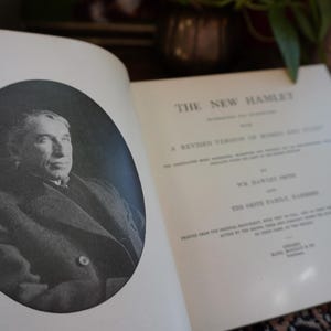 May include: An open book with the title "THE NEW HAMLET" and a black and white portrait of a man in an oval frame. The book's pages are off-white, and the text is in black.