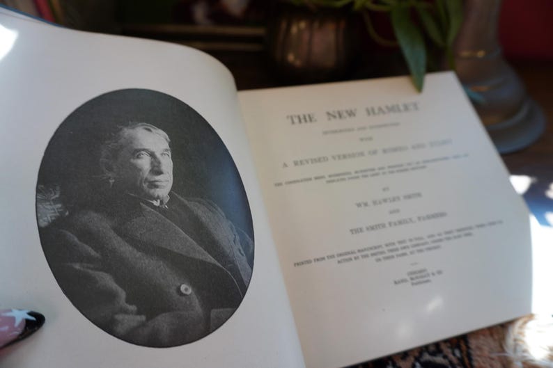 May include: An open book with the title "THE NEW HAMLET" and a black and white portrait of a man in an oval frame. The book's pages are off-white, and the text is in black.