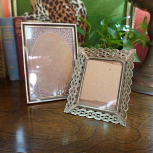 May include: Two vintage picture frames. One is rectangular with a gold border and an oval interior. The other is a smaller, ornate, silver-coloured frame with a decorative chain-link design. Both frames are empty.